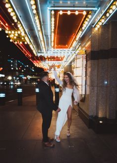 a man and woman walking down a street at night with lights on the buildings behind them