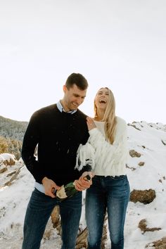 a man and woman standing in the snow holding wine bottles, laughing at each other