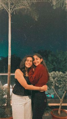 two women standing next to each other under a palm tree at night with their arms around each other