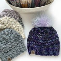 several knitted hats next to a white bowl with a purple pom - pom