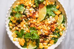 a white bowl filled with corn, cilantro and lime wedges on top of a table