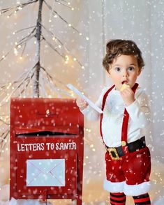 a little boy dressed as santa clause standing in front of a mailbox and christmas tree