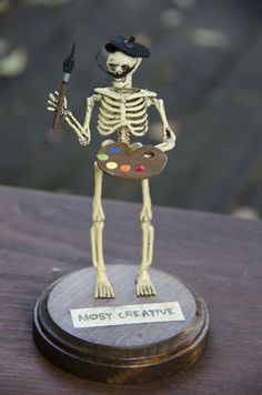 a skeleton holding a paintbrush and an artist's palette on a wooden table