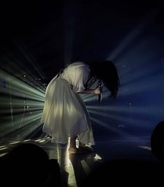 a woman in a white dress standing on stage with her hands over her head and lights behind her