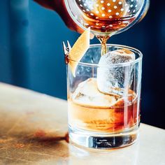 someone is pouring something into a glass with ice and an orange peel in it on the table