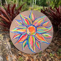 a colorful sunflower painted on top of a wooden plaque surrounded by leaves and plants