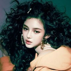 a young woman with long curly hair wearing gold earrings and an orange top is posing for the camera