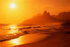 the sun is setting over an ocean with mountains in the background and people walking on the beach