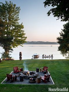 a fire pit surrounded by red chairs in the middle of a grassy area next to a lake