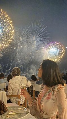 a woman sitting at a table with a glass of wine in front of fireworks