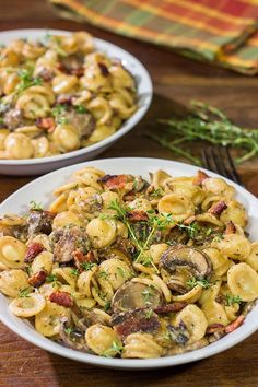 two white bowls filled with pasta and mushrooms on top of a wooden table next to a fork