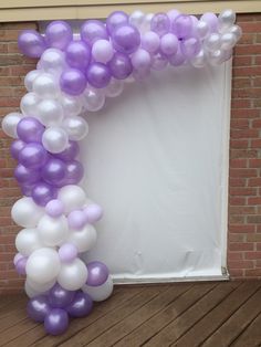 a purple and white balloon arch on a wooden floor in front of a brick wall