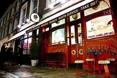 the outside of a restaurant at night with people standing outside and looking in through the windows