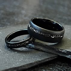 two black wedding bands sitting on top of each other in front of a stone slab