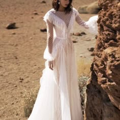 a woman in a long white dress standing next to a large rock on the desert