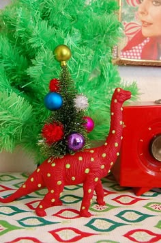 a red toy dinosaur next to a green christmas tree with ornaments on it's head