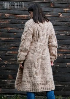 a woman standing in front of a wooden wall wearing a long cable knit coat and jeans