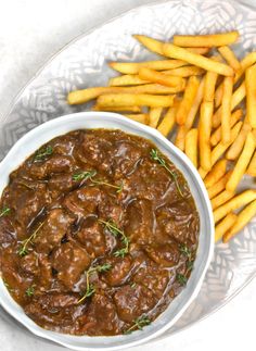 a white bowl filled with meat and fries on top of a glass plate next to a pile of french fries