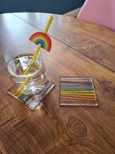 a wooden table topped with a glass filled with water and rainbow straws next to coasters