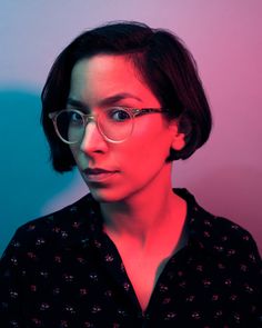 a woman with glasses is standing in front of a blue and pink background, looking at the camera