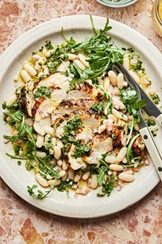 a white plate topped with chicken, beans and greens next to a knife and fork