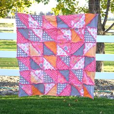 a patchwork quilt hanging from a tree in front of a white fence and green grass