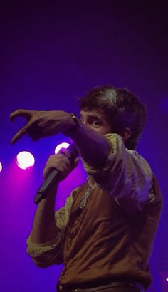 a man standing in front of a microphone on stage with purple lights behind him and his hand up to his mouth