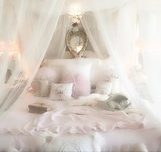 a white bed covered in lots of pink and white pillows next to a window with sheer curtains