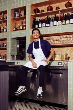 a man sitting on top of an oven in a kitchen next to shelves filled with bottles