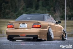 the rear end of a tan car parked in a parking lot with trees in the background