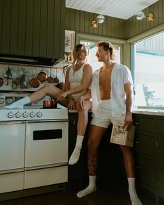 a man and woman sitting on the counter in a kitchen with an oven, stove top and cabinets