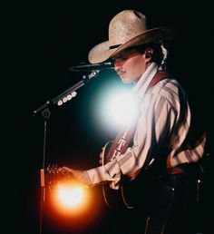 a man wearing a cowboy hat and holding a guitar in front of a microphone on stage