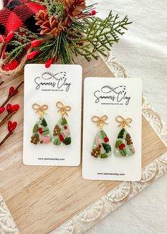 two small christmas tree earrings on top of a wooden table next to pine cones and red berries