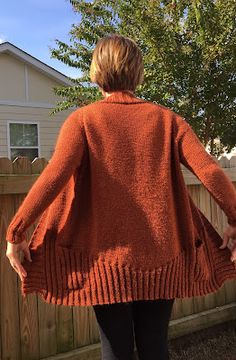 a woman standing in front of a fence with her back to the camera, wearing an orange sweater