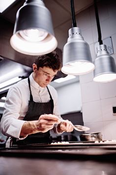 a man in an industrial kitchen preparing food on a plate and lights hanging from the ceiling above him