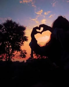 a woman making a heart shape with her hands in front of the sun at sunset