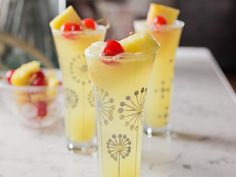 three glasses filled with yellow drinks sitting on top of a white marble counter topped with pineapple slices and cherries