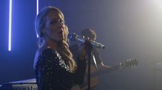 a woman singing into a microphone while holding a guitar in front of her and another person behind her