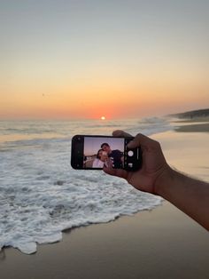 a person holding up a cell phone to take a photo on the beach at sunset