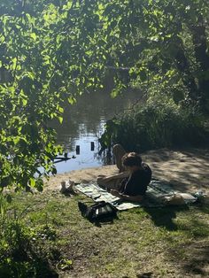 a woman sitting on top of a blanket next to a body of water with trees in the background