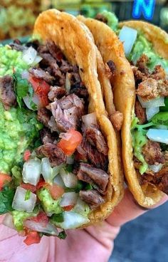 two tacos with meat, lettuce and tomatoes on them are held in someone's hand