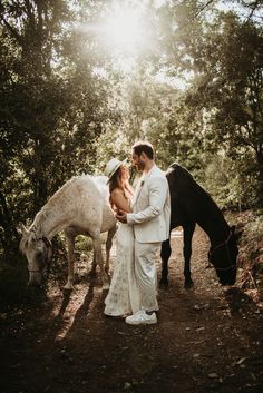 a bride and groom standing next to horses in the woods with sunlight shining through trees