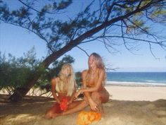 two women in bathing suits sitting on the sand under a tree near the ocean and beach