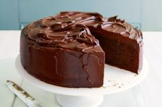 a chocolate cake with one slice missing from it on a white cake platter next to a knife and fork