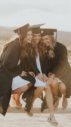 three women in graduation gowns and caps sitting on a fence with their arms around each other