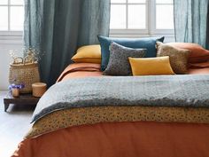 a bed with many pillows on it in front of a window and blue drapes