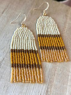 two tone beaded earrings with gold and white accents on a wooden table next to a bottle