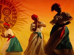 three women in colorful dresses are dancing on stage with an orange and yellow backdrop behind them