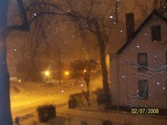 the snow is falling down in front of a house and trees on a snowy night