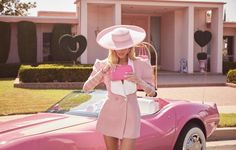 a woman in a pink dress and hat standing next to a pink sports car with the hood up